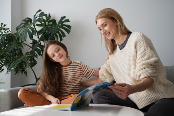 Woman and girl looking at a book together, smiling. Sitting on a couch in a bright room.