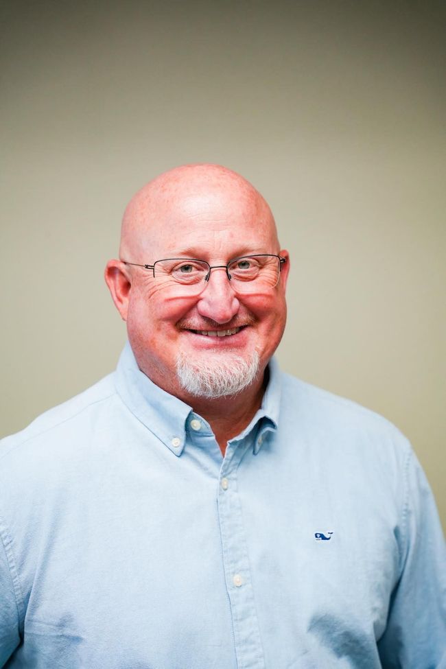 Bald man with glasses, smiling, wearing a light blue button-down shirt with a small whale logo, indoors.