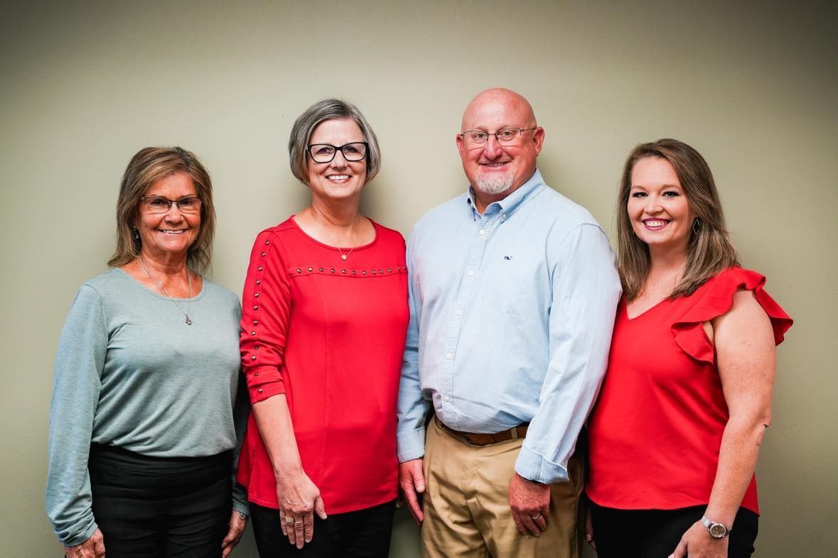 Four people standing in front of a neutral wall; two women in red tops, two others in blue/grey shirts, smiling.
