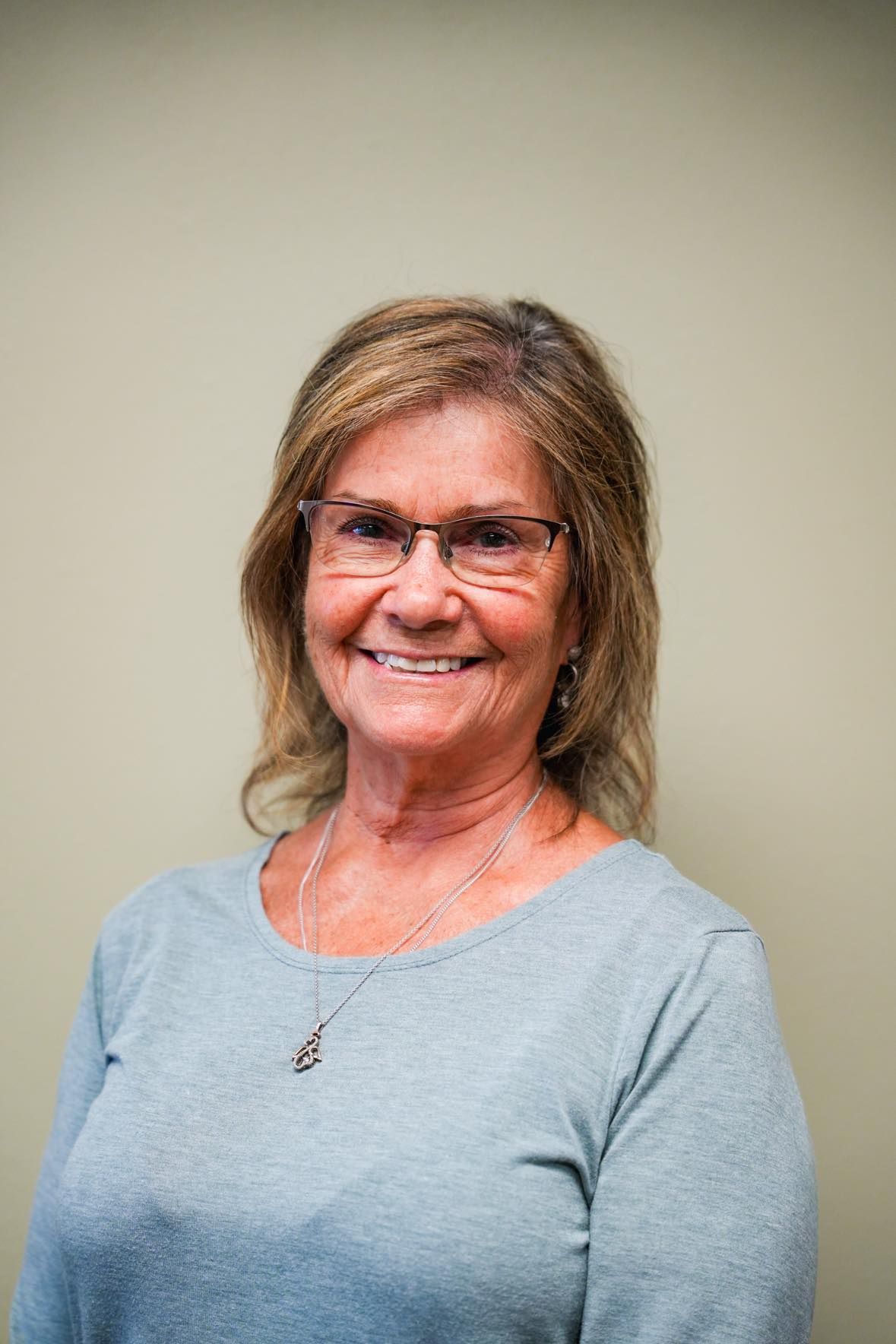 Smiling older woman with glasses wearing a gray shirt, standing in front of a neutral wall.