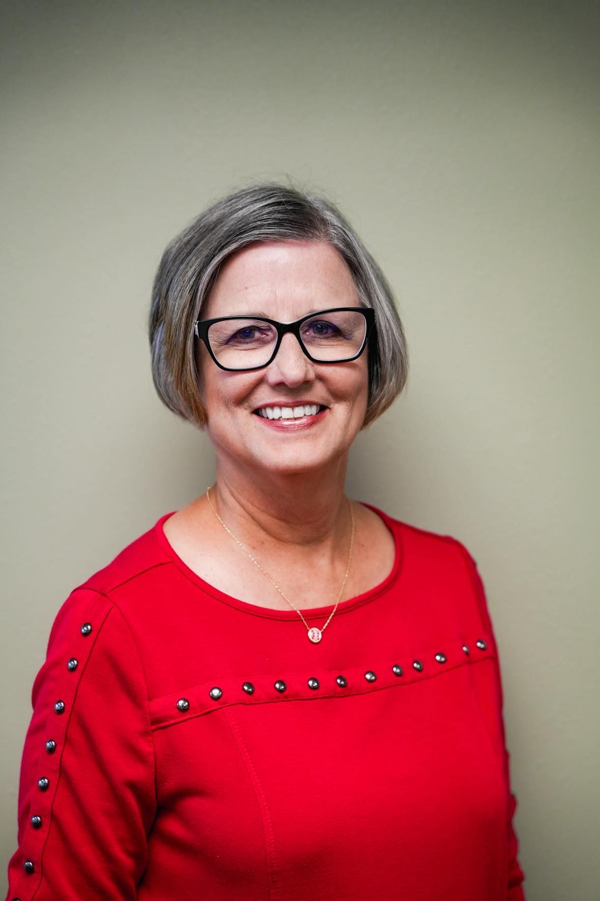 Woman with gray hair and glasses wearing a red sweater, smiling, in front of a light green wall.