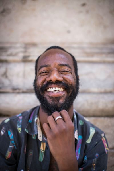 A Black man with a full beard smiles broadly, hand to his chin, in front of a stone structure.