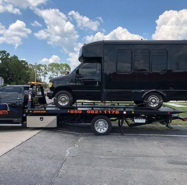 Black shuttle bus on a flatbed tow truck in a parking lot under a partly cloudy sky