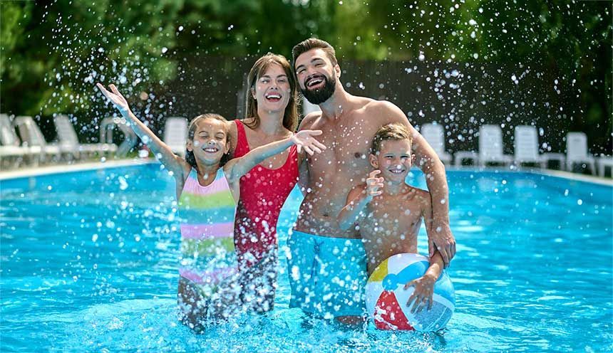 Family smiling and splashing in a pool. Daughter with arms up, son holding ball. Blue water, sunny day.