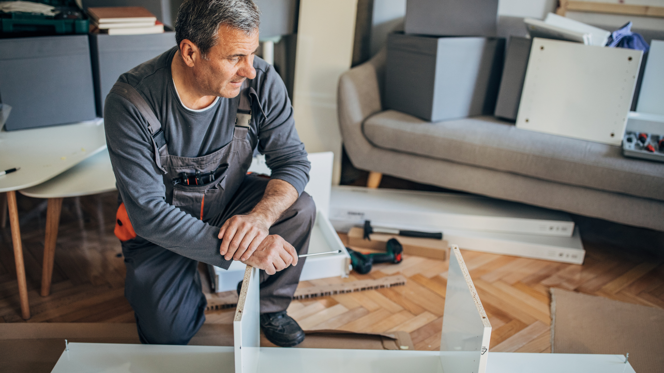 A man is kneeling down in a living room while assembling a piece of furniture.