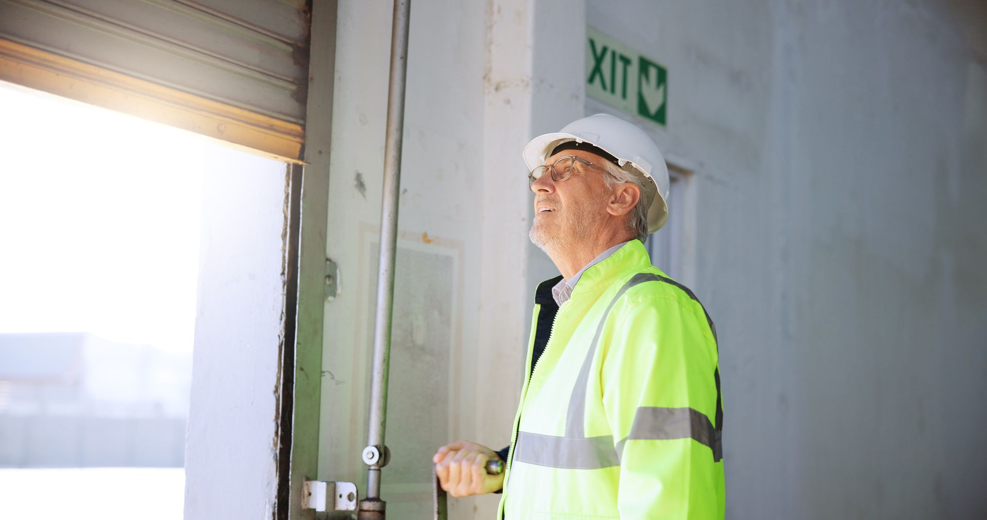 Worker in hard hat and reflective jacket inspecting an industrial doorway beneath an exit sign. Worker in hard hat and reflective jacket inspecting an industrial doorway beneath an exit sign.