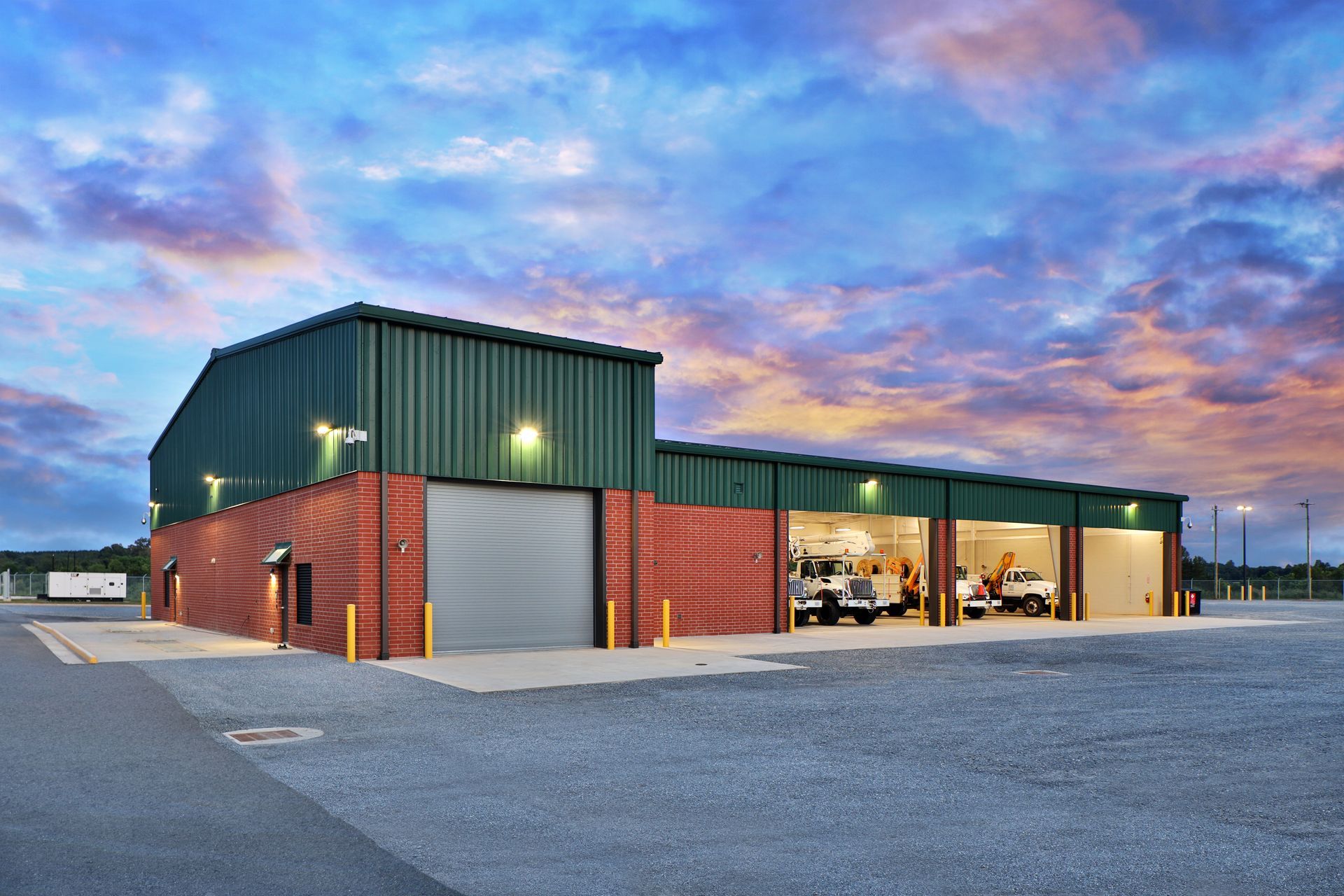 Green and red brick building with three open garage bays, vehicles inside, and a sunset.