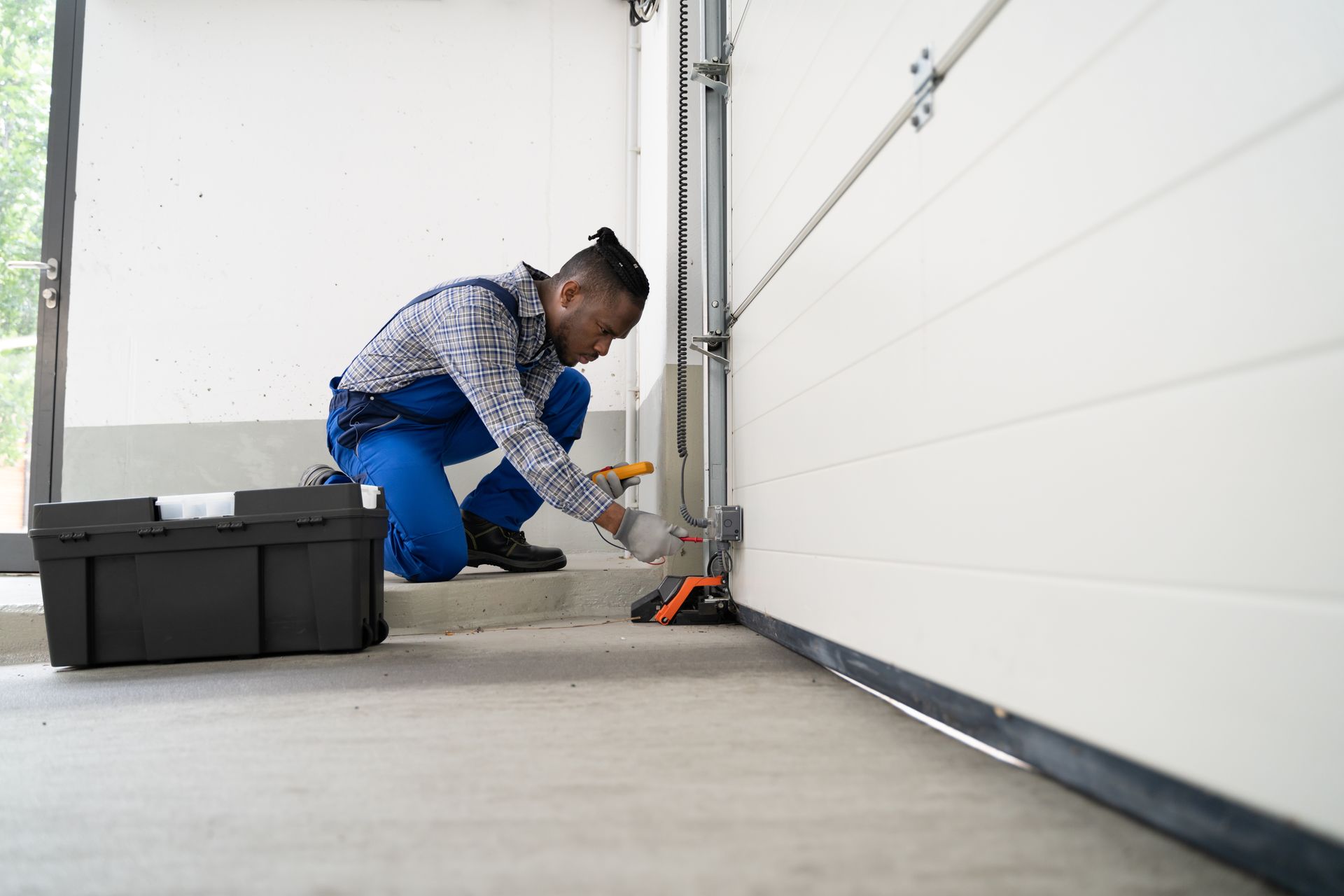 Man in blue overalls repairs garage door, using tools from a black toolbox.
