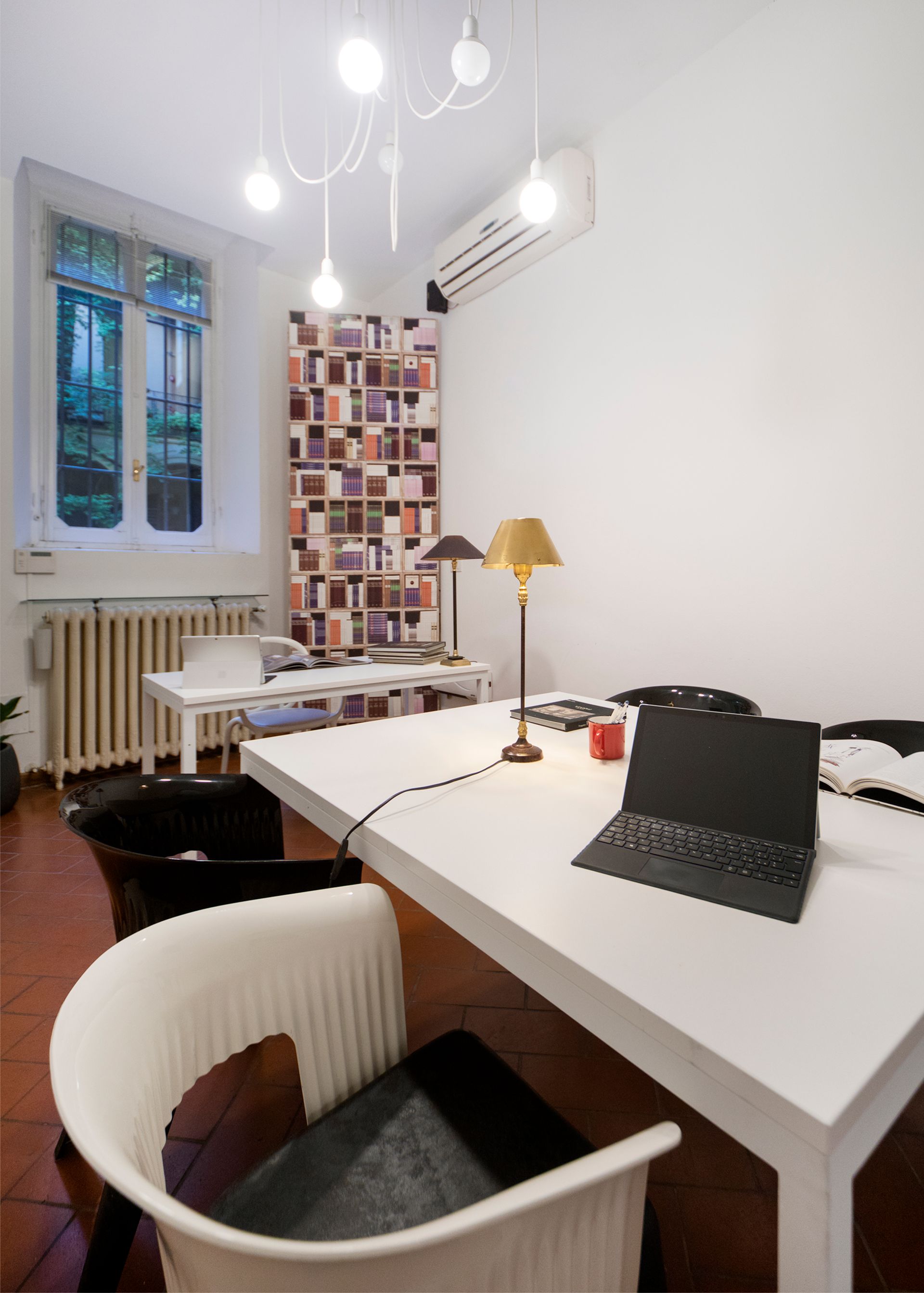 A well-lit home office with white desks, chairs, and a bookshelf; window, air conditioner, and overhead lights present.