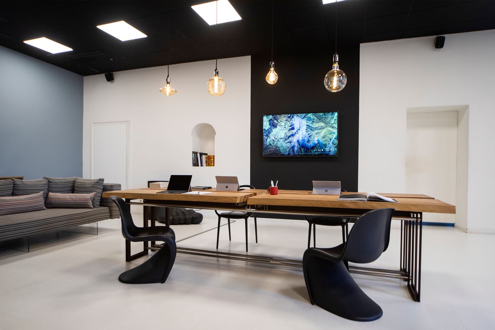 Modern office space with a long wooden table, chairs, and a mounted TV on a black wall.