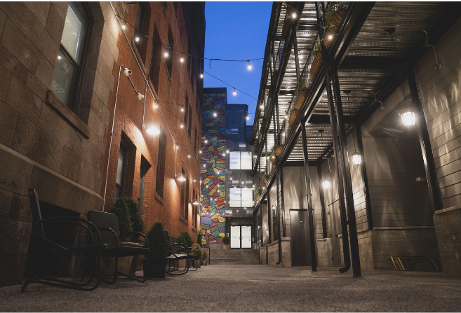 A narrow alley at dusk with string lights, benches, and a colorful mural.