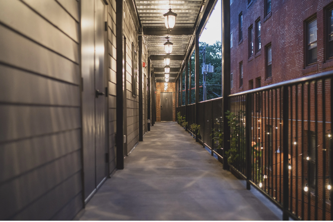 A hallway with lamps, leading to a brick building. Railing on the right and doors on the left.
