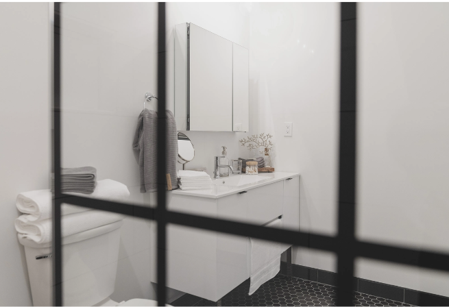 White bathroom with black accents, seen through a black paneled window. Toilet, sink, and mirror are visible.