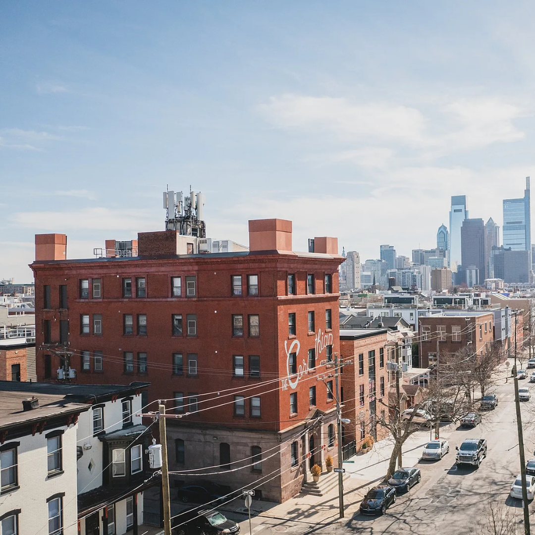 Brick buildings line a street with cars, with a city skyline visible in the distance on a sunny day.