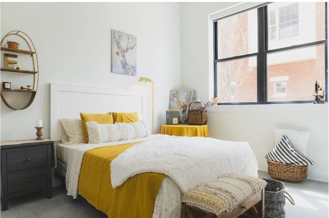 Bedroom with a white bed, mustard-yellow accents, and a large window letting in light.