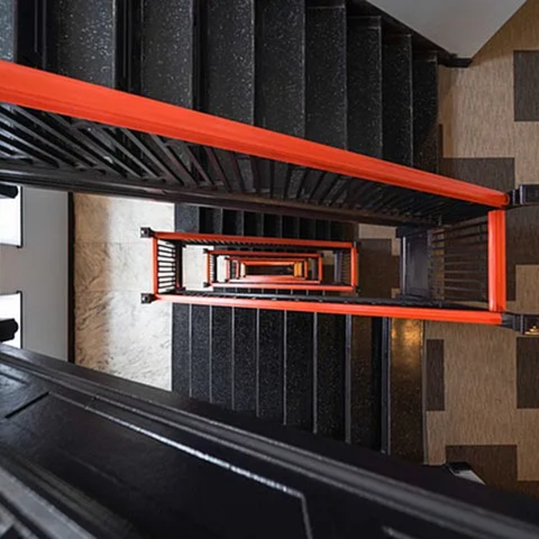 Looking down a stairwell with black steps, orange handrails, and patterned beige flooring.