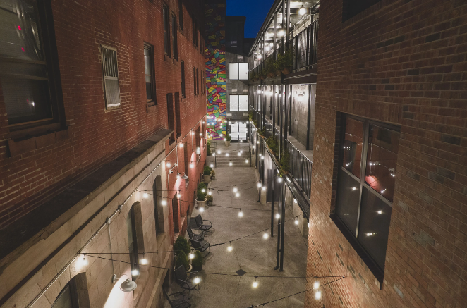Narrow alleyway at dusk, brick buildings, string lights, colorful art in the distance.