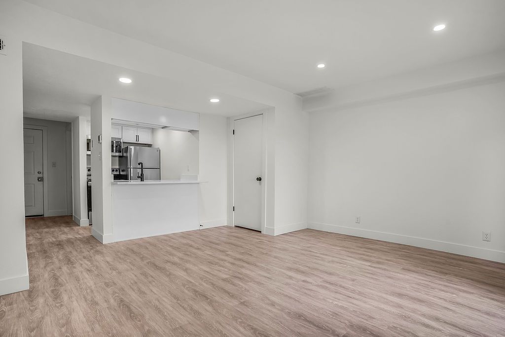 Empty living space with light wood-look flooring, white walls, and a small kitchen opening.