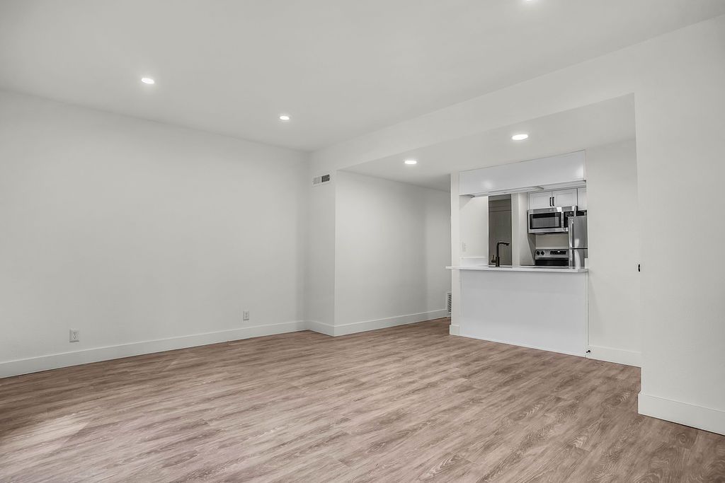 Empty room with light brown carpet, white walls and a pass-through to a kitchen area.