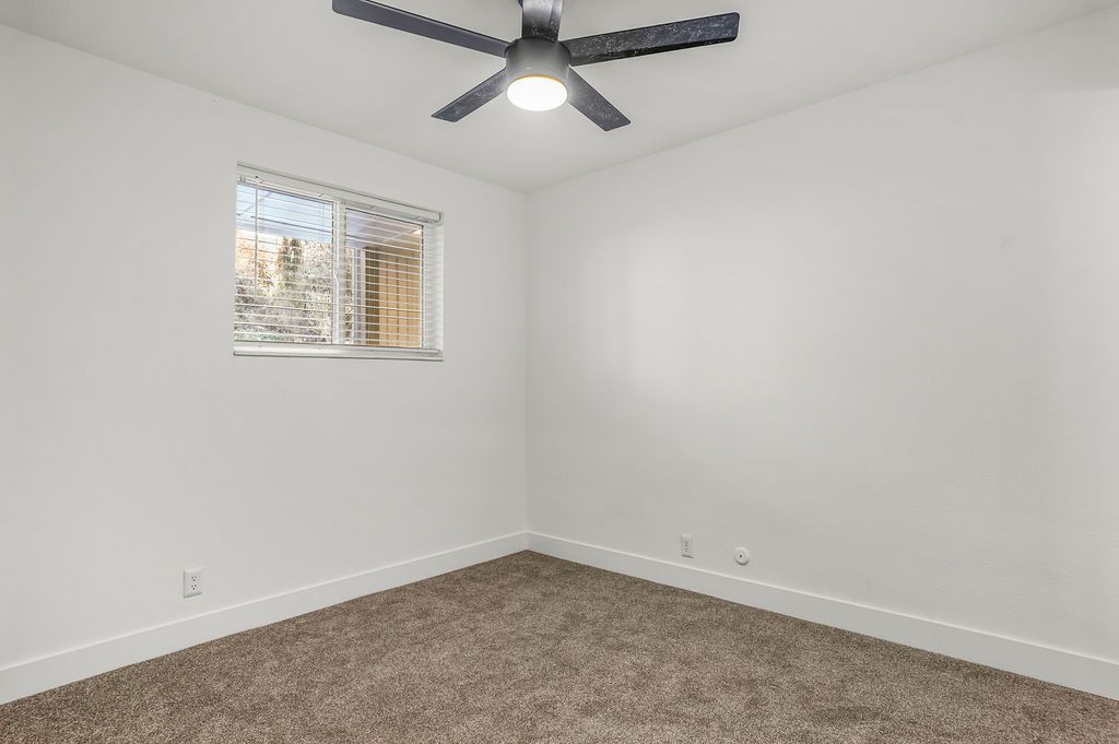 Empty bedroom with brown carpet, white walls, window, and ceiling fan.