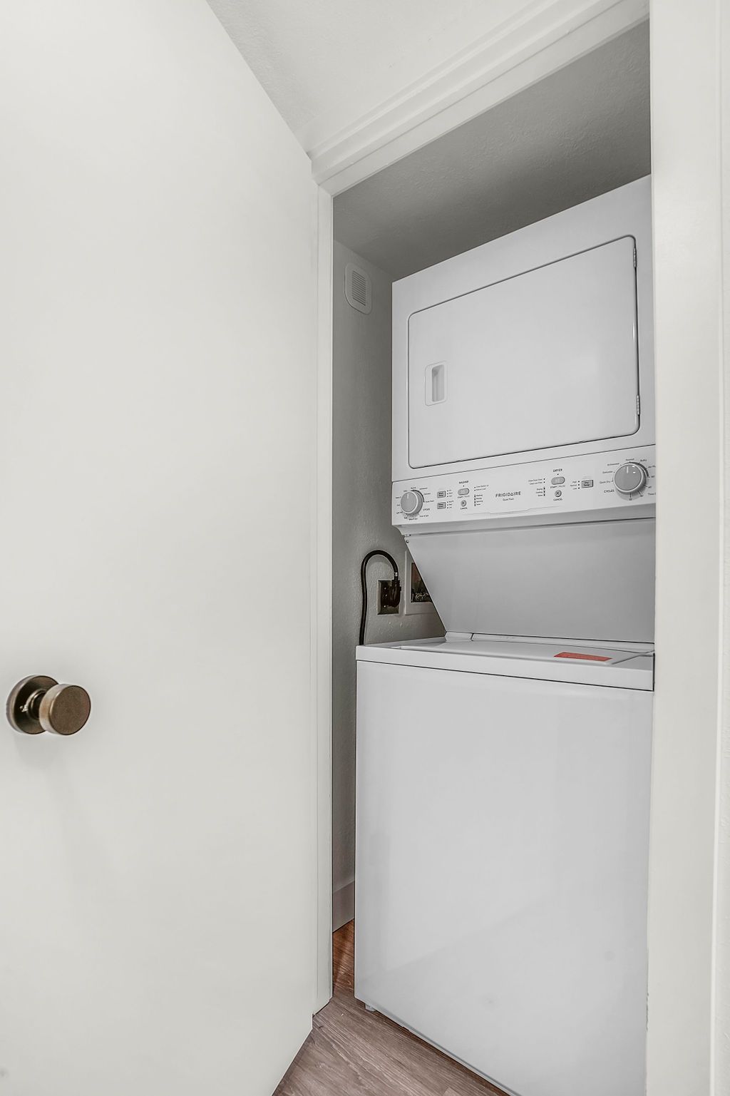White stacked washer and dryer unit in a small, white-walled laundry closet; door is partially open.