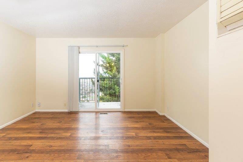 Empty room with wood floors and sliding glass door leading to balcony overlooking trees.