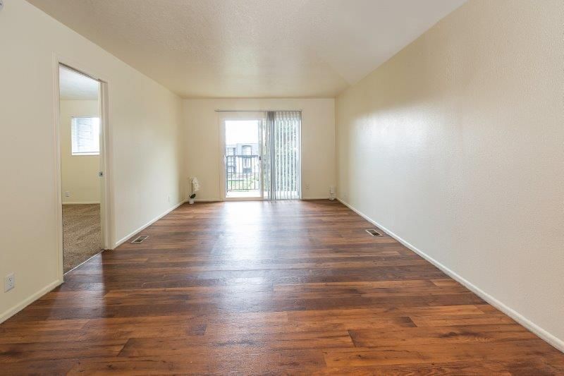 Empty living room with brown wood floor, sliding glass door, and cream-colored walls.