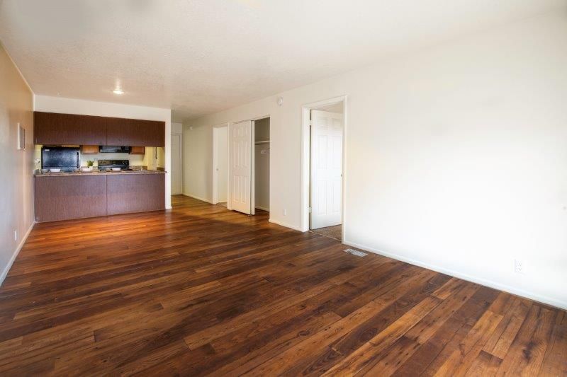 Empty apartment interior with dark hardwood floors, white walls, and a small kitchen.