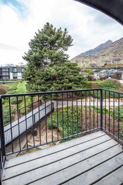 Balcony overlooking a bridge, tree, and distant buildings. Black railing, grey wood floor, cloudy sky.