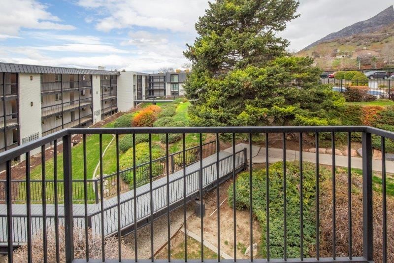 Black metal balcony overlooking a courtyard with a bridge, green foliage, and apartment buildings, with a mountain in the background.