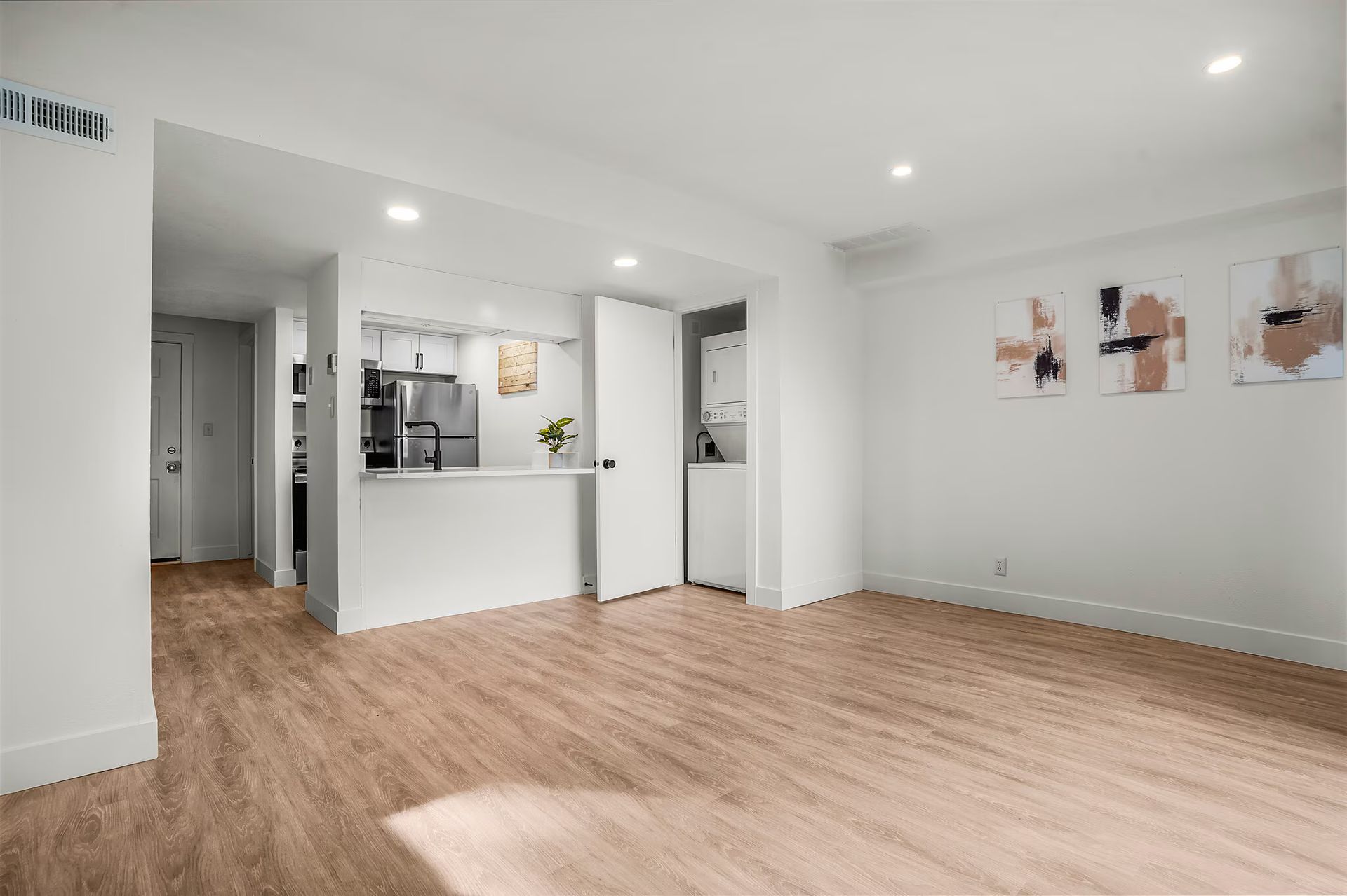 Empty apartment interior with light wood flooring, white walls, open kitchen, and abstract art.