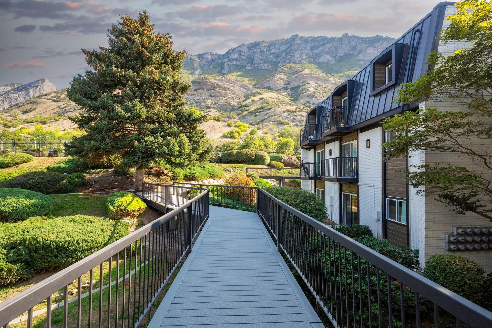 A wooden walkway with black railings leads to a building with a backdrop of mountains and greenery.