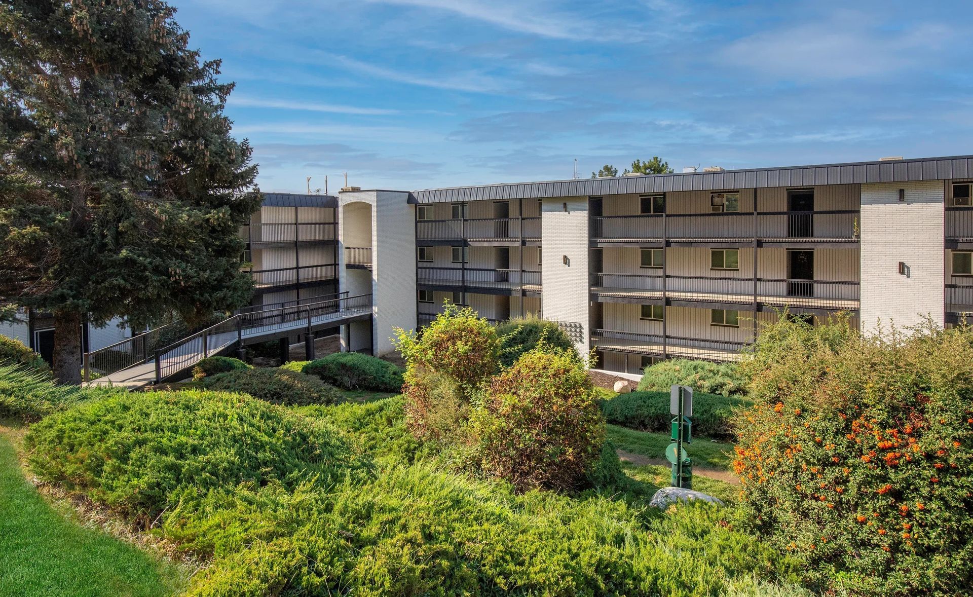 Three-story building with balconies, connected by exterior walkways, set amidst green landscaping under a blue sky.