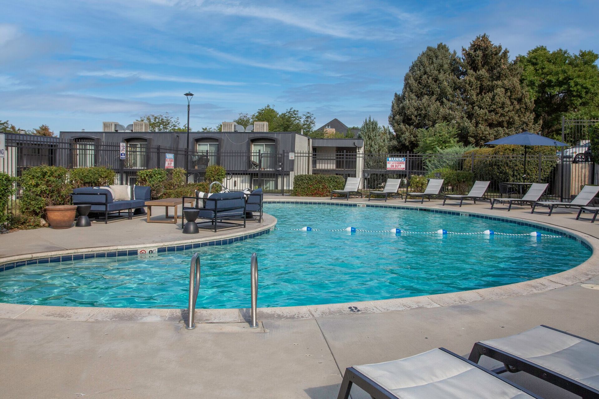 Pool with lounge chairs in front of apartments under a blue sky.