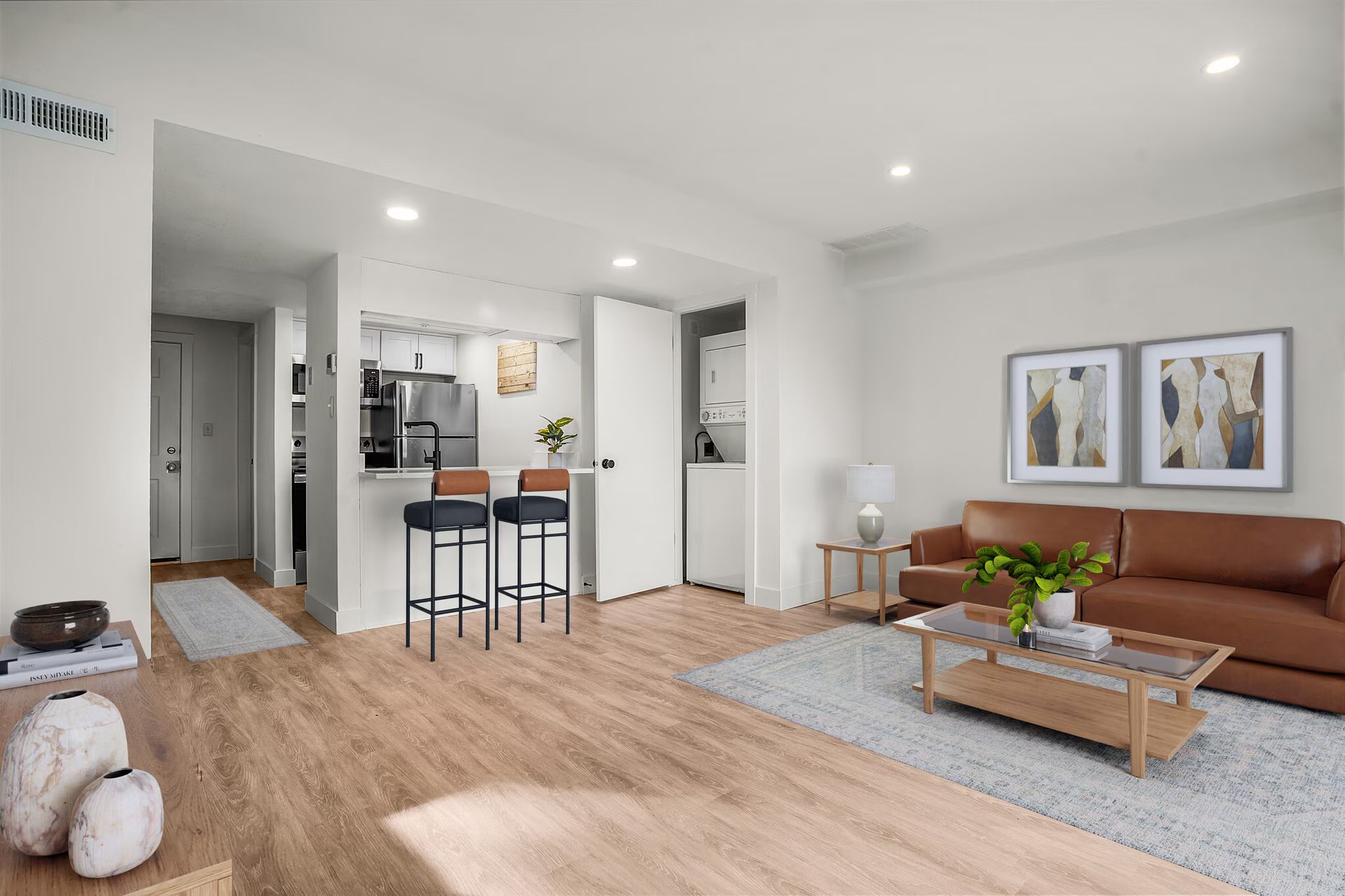 Living room and kitchen: light wood floors, brown couch, kitchen bar with stools, artwork.