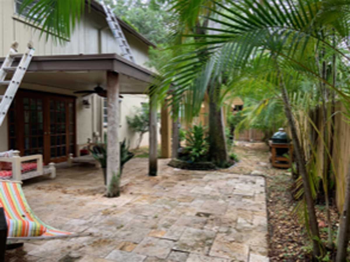 A patio with a hammock and a ladder in the backyard of a house surrounded by palm trees.