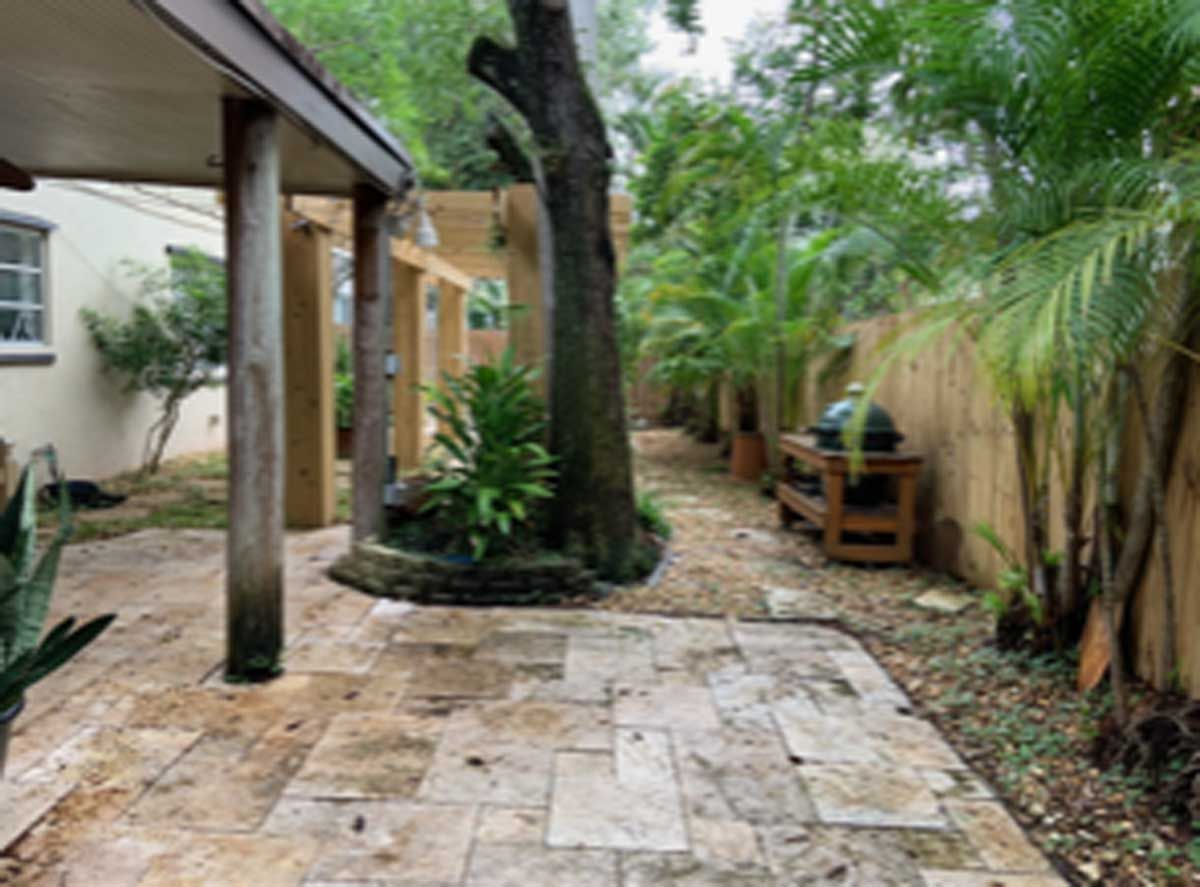 A patio with a covered walkway leading to a house surrounded by trees and bushes.