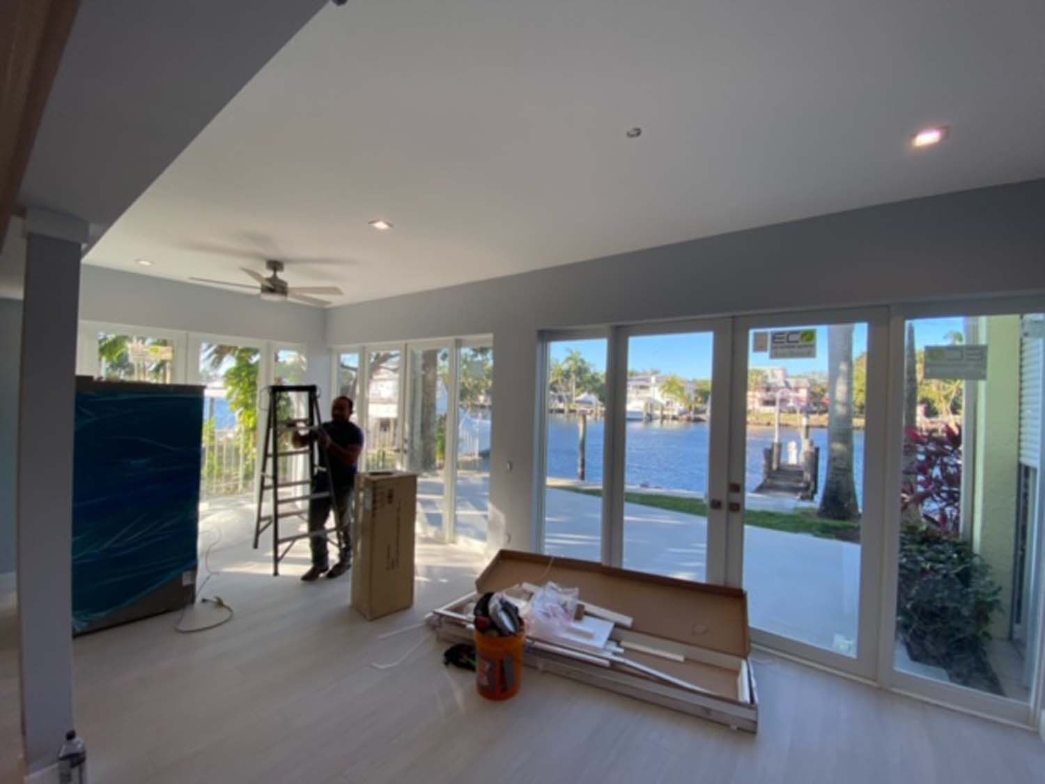A man is standing on a ladder in a living room with a lot of windows.