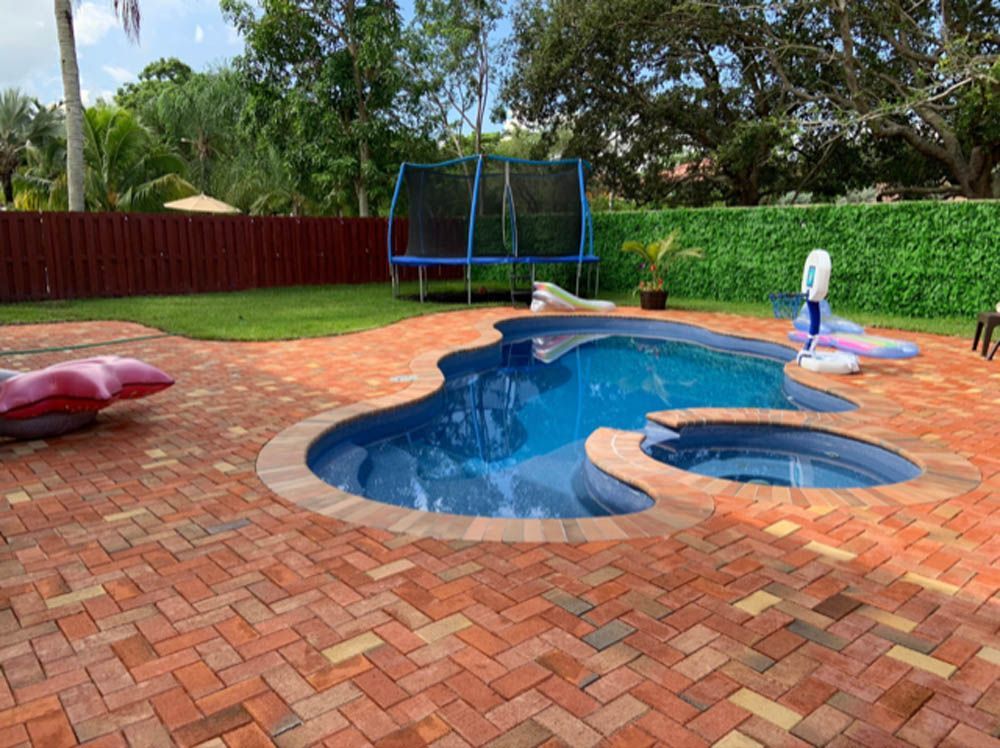 A large swimming pool is surrounded by a brick patio and a trampoline.