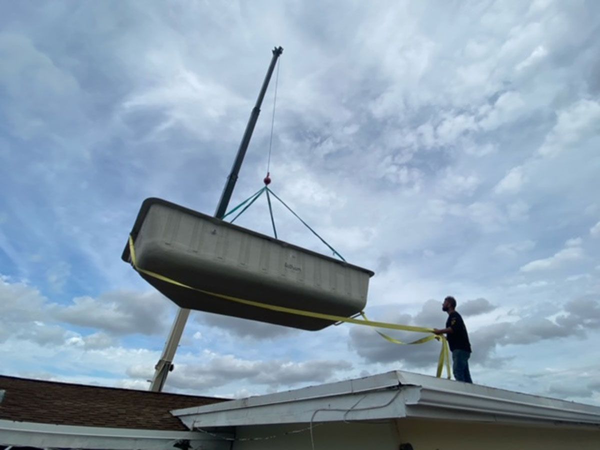 A man is standing on the roof of a house while a crane lifts a pool into the air.