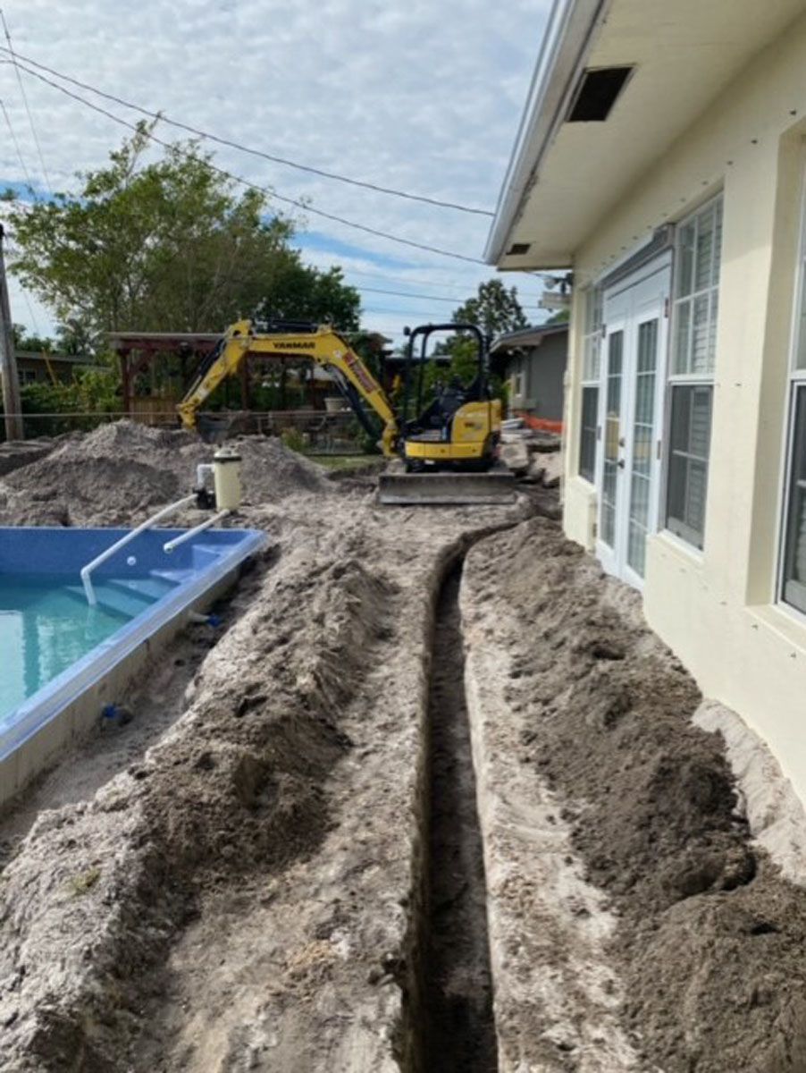 A yellow excavator is digging a trench in front of a house next to a pool.