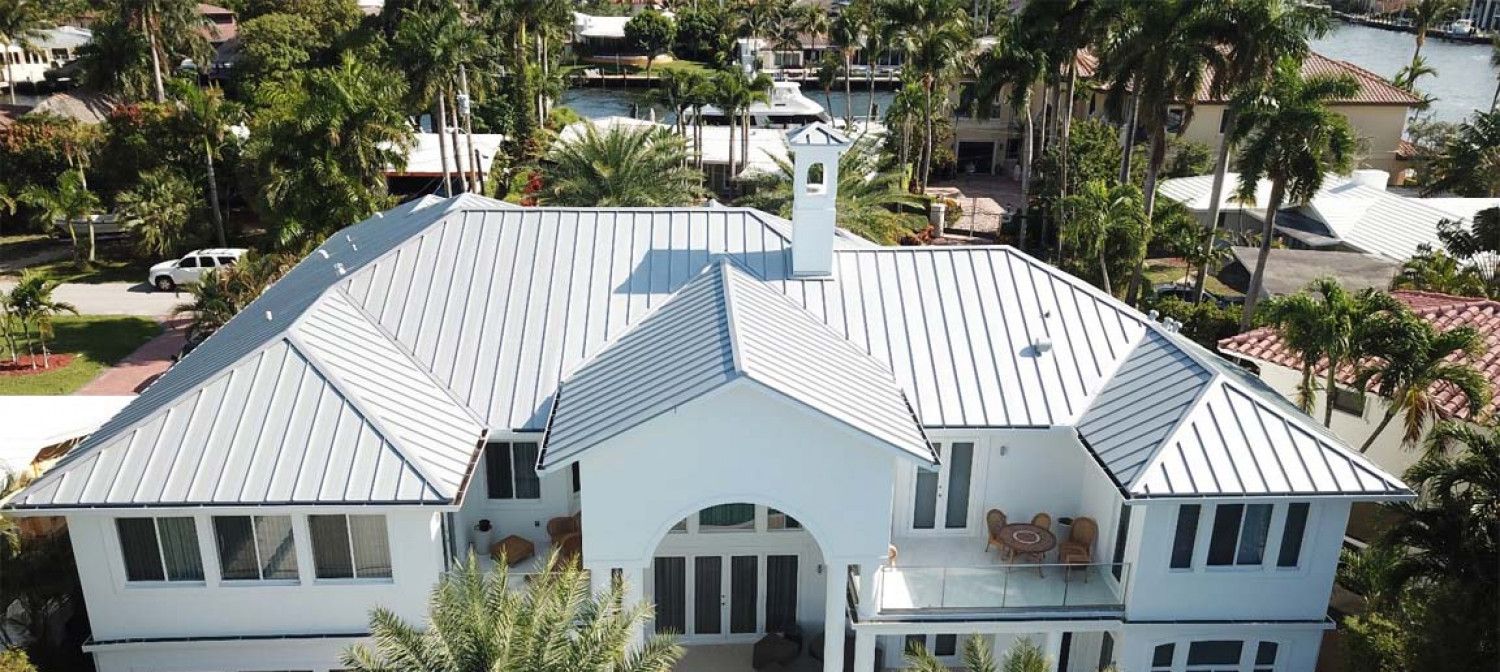 An aerial view of a large white house with a metal roof surrounded by palm trees.