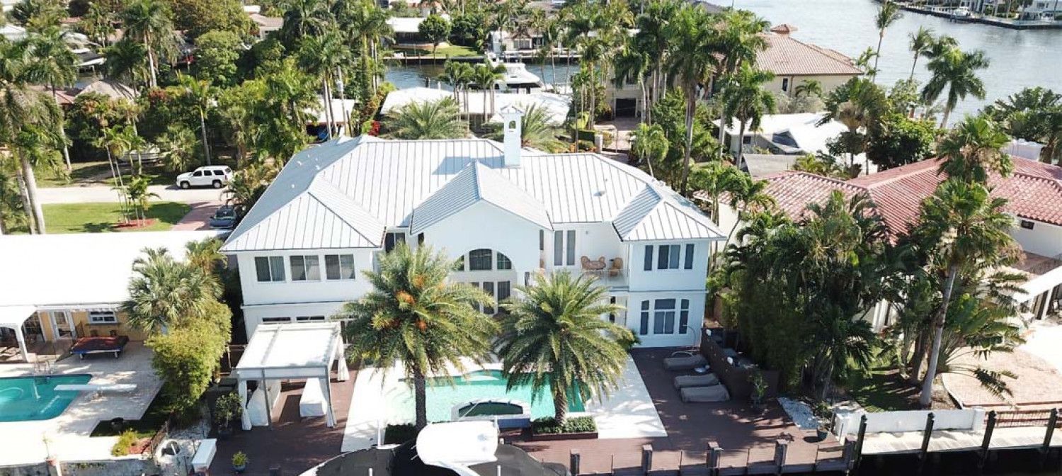 An aerial view of a large white house with a pool surrounded by palm trees.