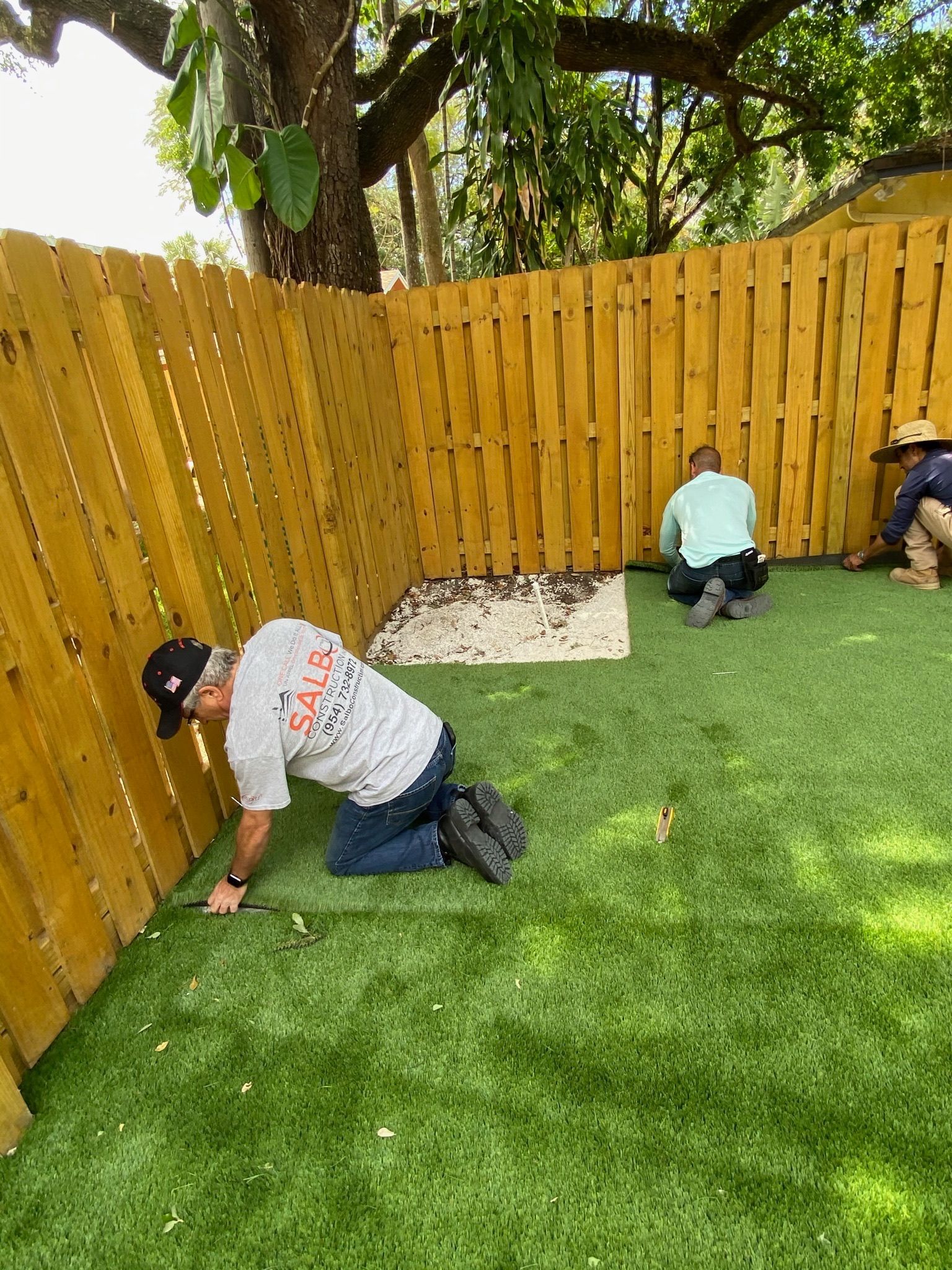 Three men are kneeling on the grass in front of a wooden fence.