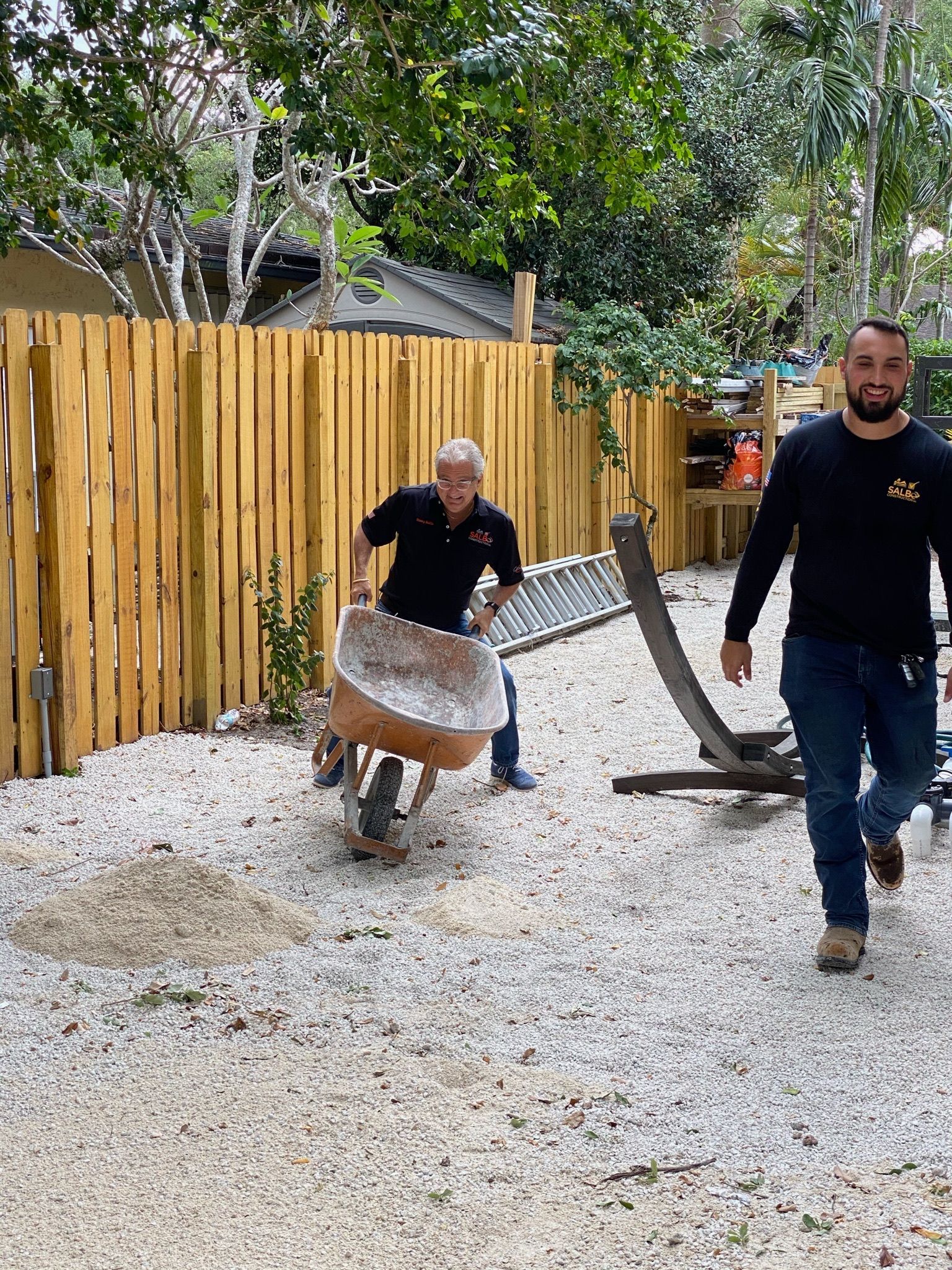 Two men are pushing a wheelbarrow down a gravel path.