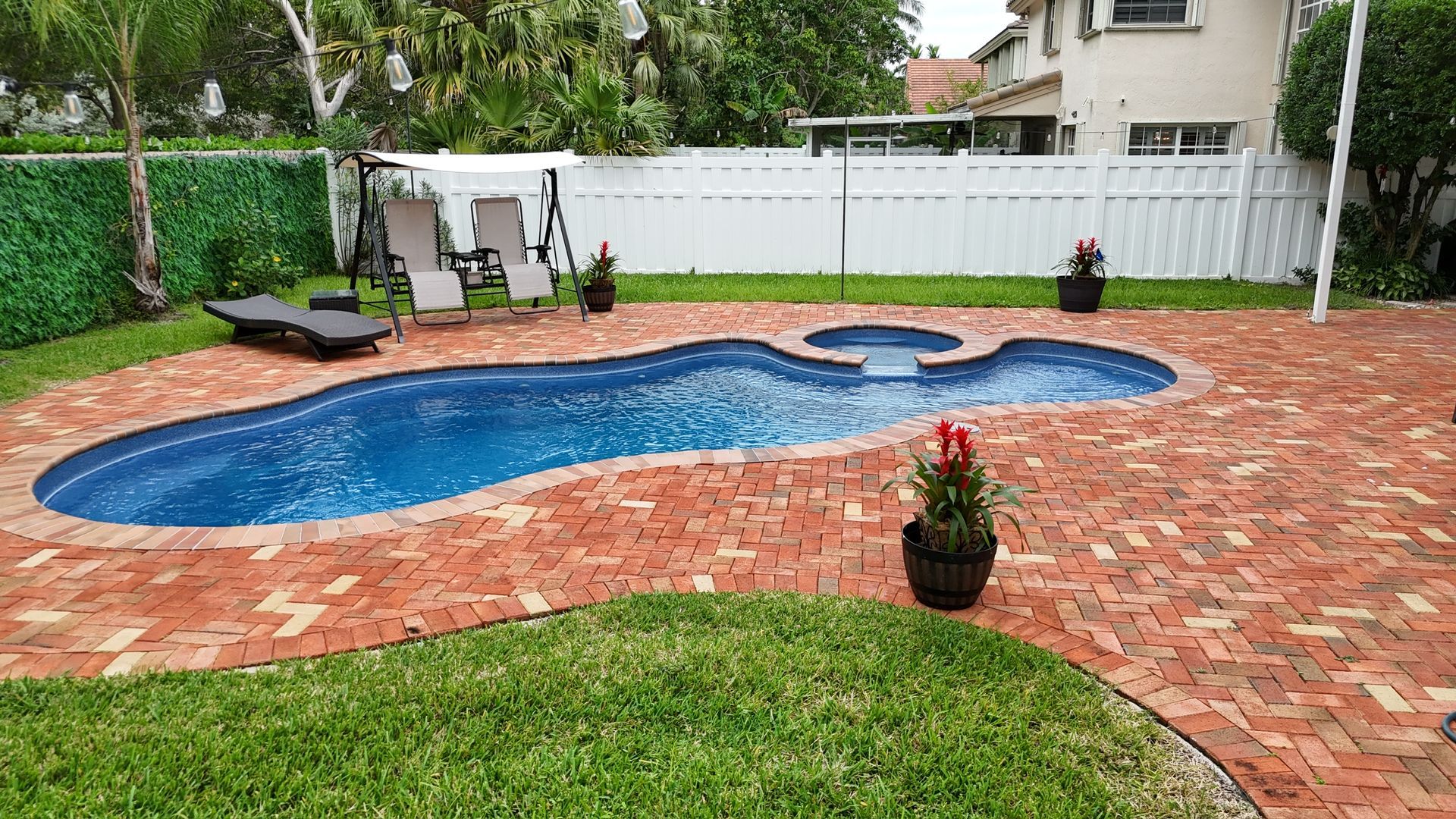 A large swimming pool is surrounded by a brick patio in a backyard.
