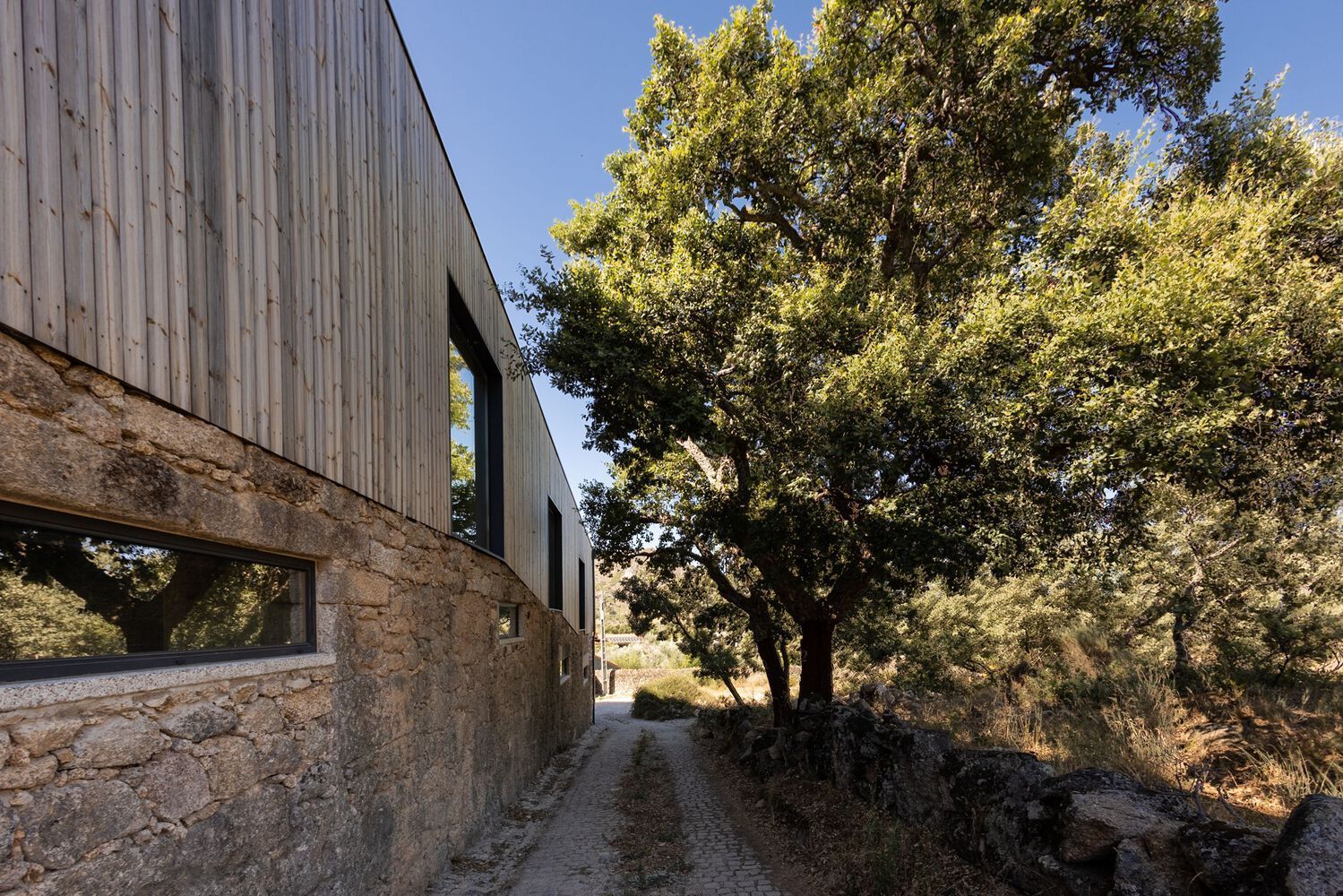 A stone building with a wooden roof is surrounded by trees and a dirt road.