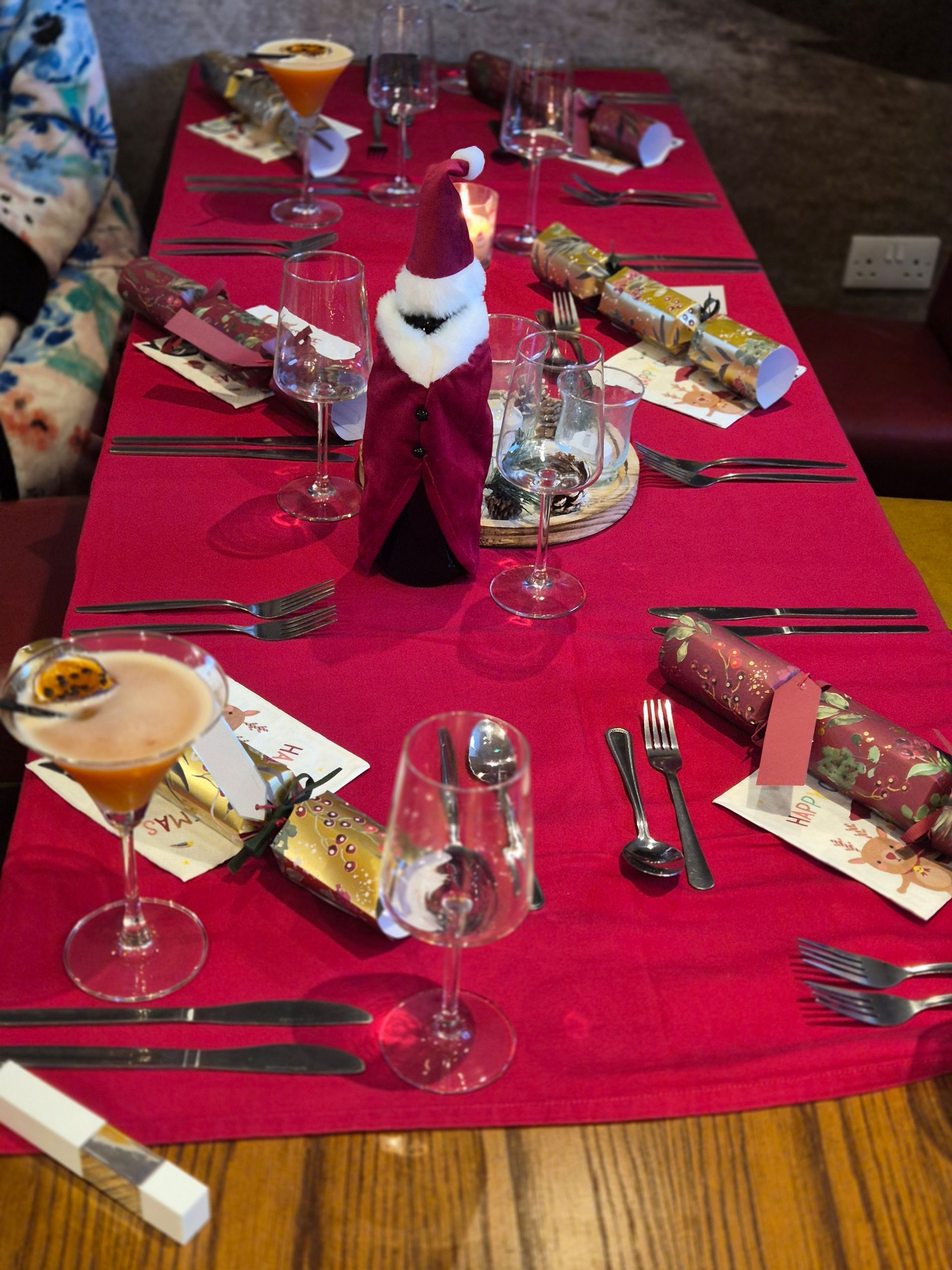 A table with six settings on a red table cover with cutlery, glasses, cocktails and Christmas crackers.