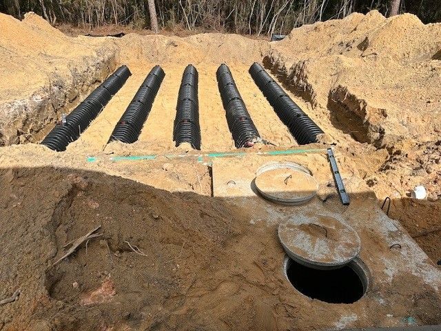 Septic system under construction: black pipes laid in trenches of tan sand. Open tank in foreground.