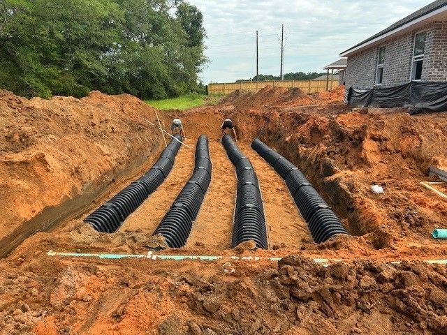 Four black corrugated pipes laid in trenches, possibly for drainage, near a house under construction.